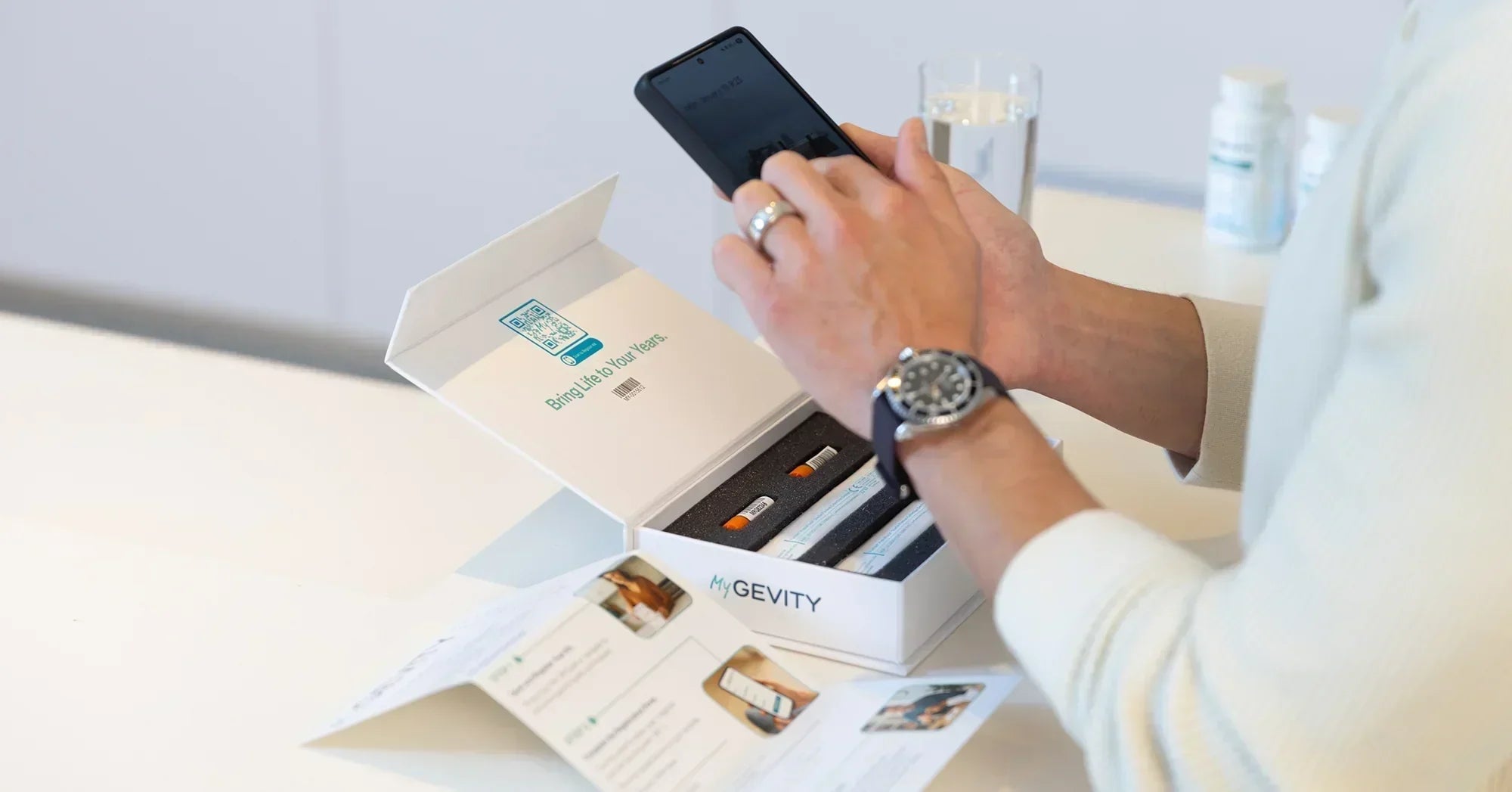 A close-up of an open MyGEVITY DNA collection kit on a white counter, featuring collection vials and a QR code for digital registration, with a person using a smartphone in the background.